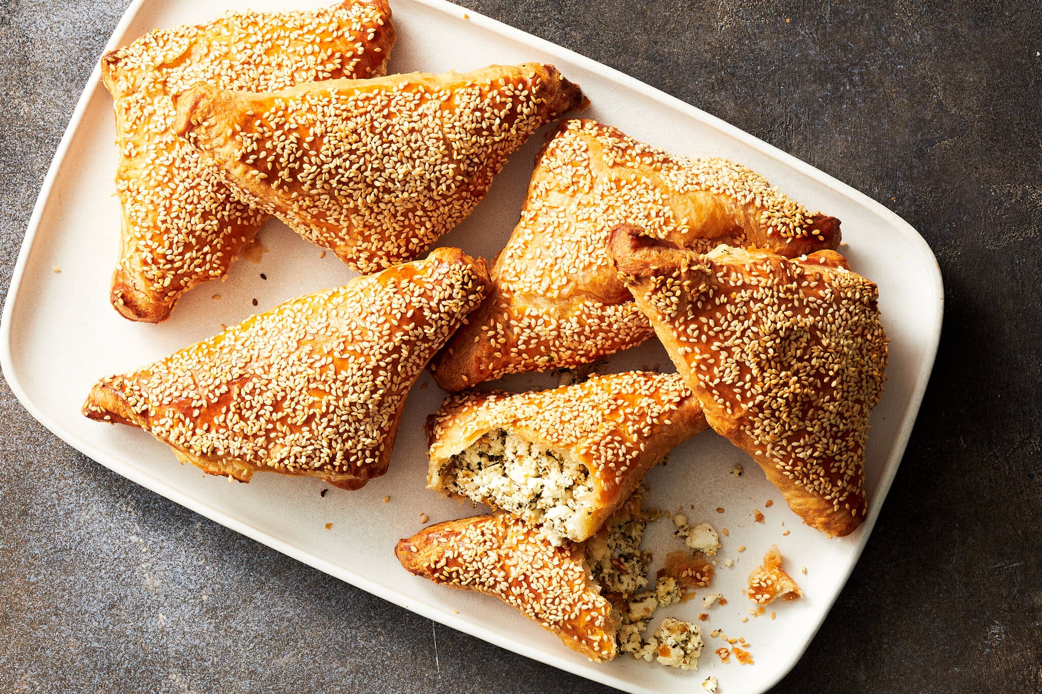 An overhead image of sesame-crusted puff pastry turnovers on a white platter. A single turnover is opened to reveal a feta filling.