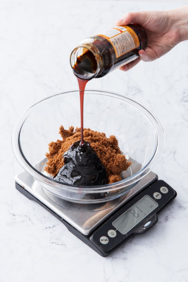 Pouring molasses into a mixing bowl sitting on a digital scale along with brown sugar.