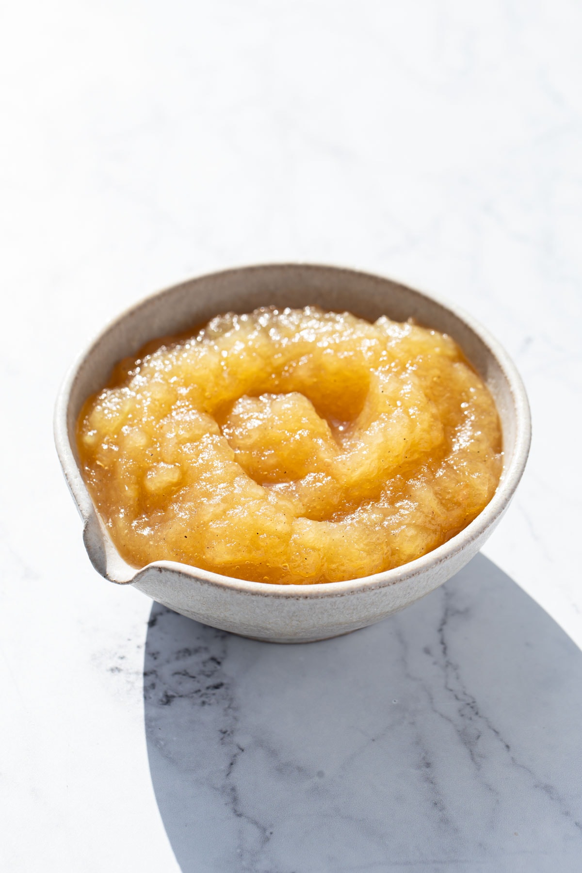 Bowl of homemade applesauce in direct sunlight on a white marble background.