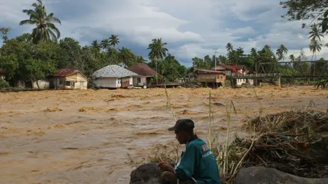 Getty Images A man sits by the side of a body of moving water on a road on Batipuh Village, West Sumatra, Indonesia, with homes submerged