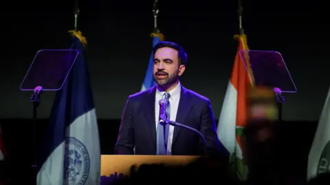 REUTERS/Jeenah Moon Zohran Mamdani speak under lights from a stage. He is wearing a blue suit with a blue tie. Flags stand behind him.