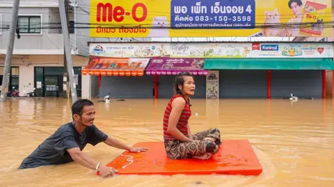 Getty Images A man transports a woman on an orange plastic board through flood waters in Hat Yai in Thailand's southern Songkhla province