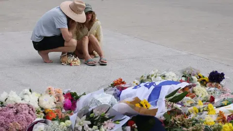 AFP via Getty Images Two women comfort each other as they stare at flowers left in tribute to the victims of Sunday's shooting attack at Bondi beach. One of the women, dressed in a yellow shirt, is sitting on the road, while the other kneels next to her with her hand on her shoulder. 