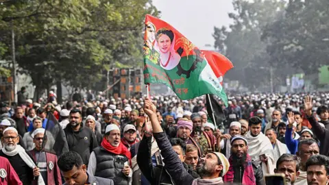 AFP via Getty Images A large crowd gathers on the street to pay their respects to Khaleda Zia. One man holds up a flag featuring her face near the front of the crowd.