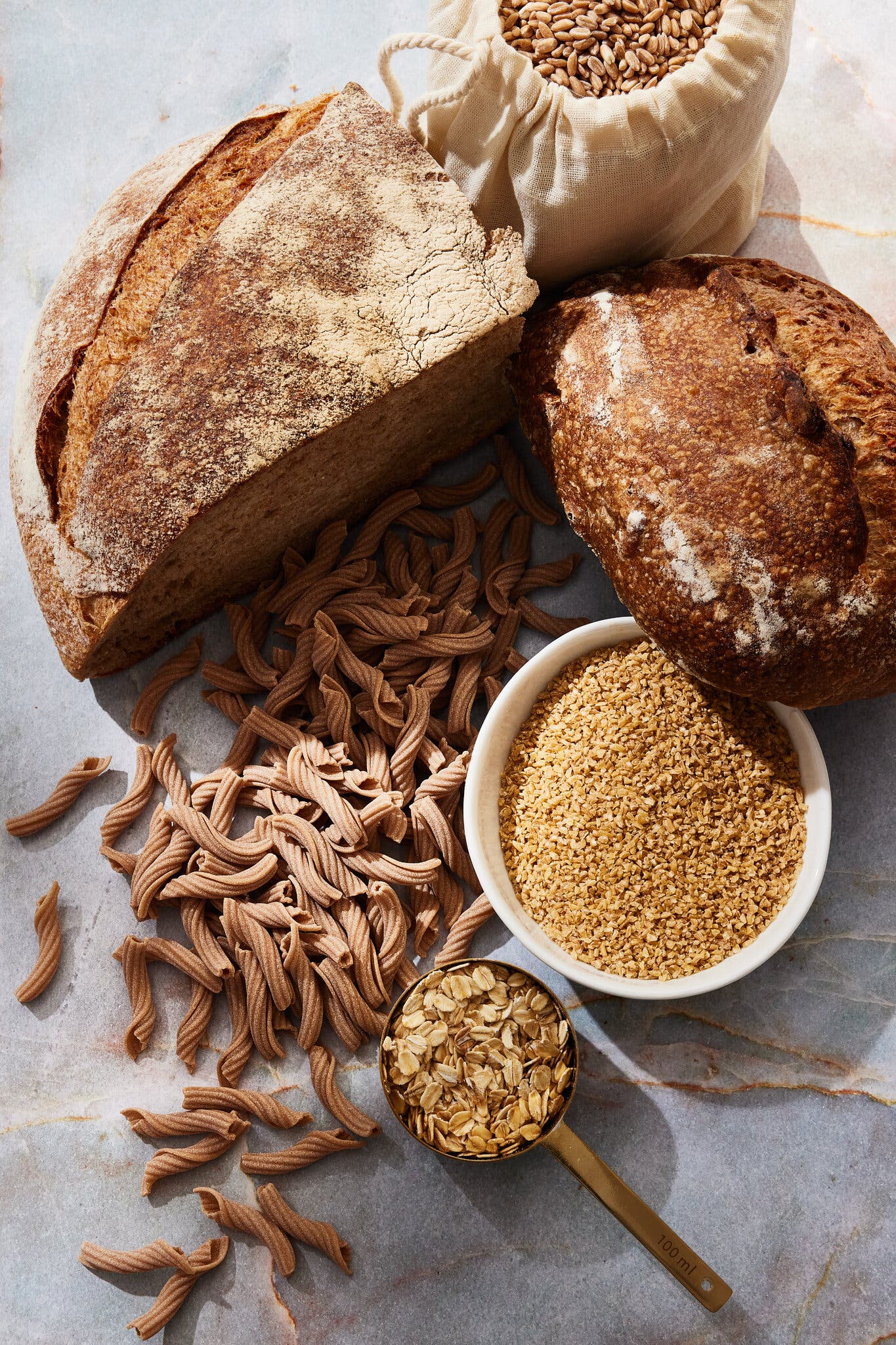 A hunk of sourdough, whole grain pasta and other grains photographed against a pale surface.
