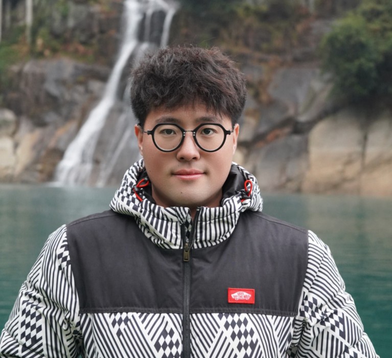 Dezhou Cao wearing a thick winter coat and standing in front of a waterfall.