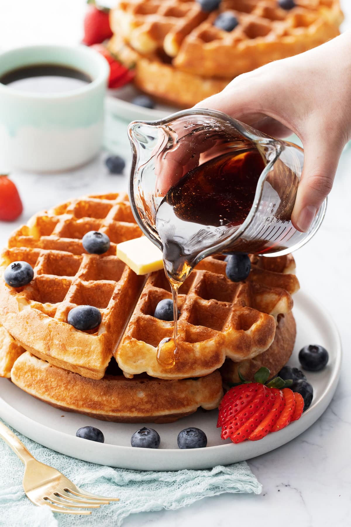 Syrup being poured over a stack of golden brown Overnight Sourdough Discard Waffles topped with berries and a pat of butter.